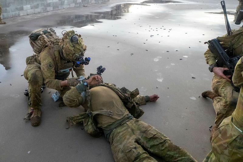 A man in uniform lies on wet concrete surrounded by spent shell casings while another kneels beside him.