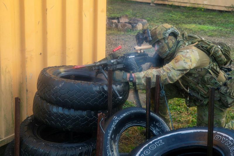 A man in uniform with camouflage face paint fires a weapon from behind a stack of tyres.