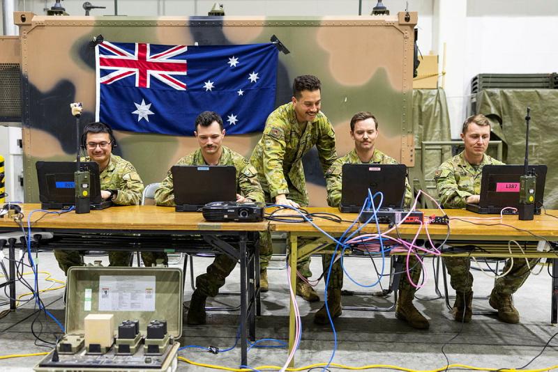 Four men in uniform sit at two tables looking at laptop computers, with another man in uniform standing in the middle, looking at one of the computers.