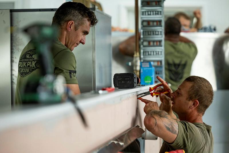 A man in uniform puts pliers into a small hole in a wall while another man in uniform stands on the other side of the wall.