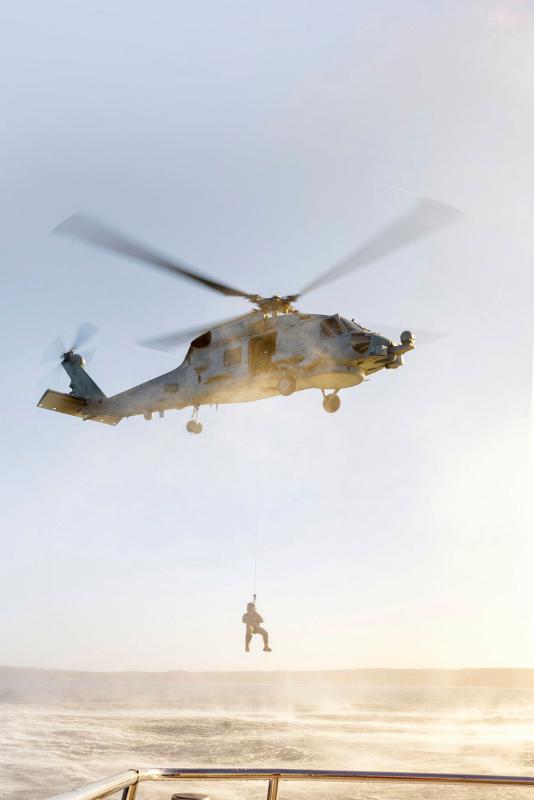 A helicopter hovers over water with a person suspended from a winch underneath.