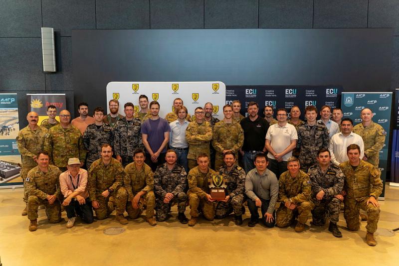 A group of military personnel group together for a photo, with two members holding a trophy.
