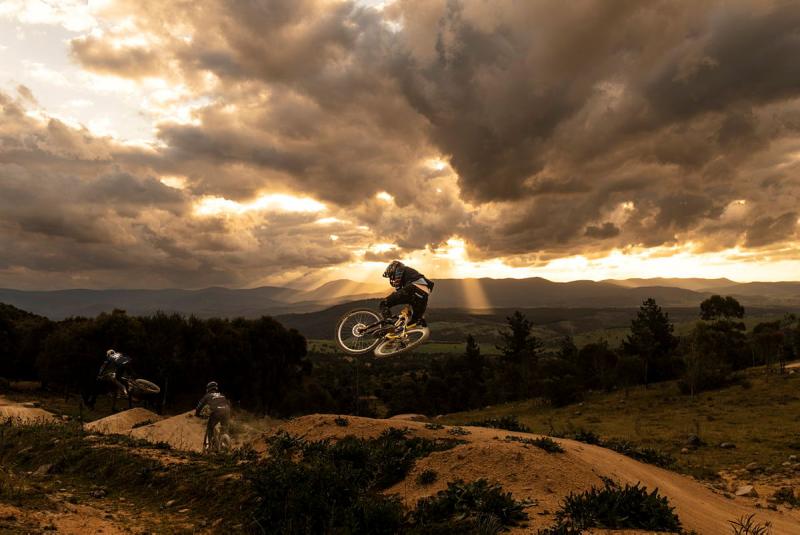 Cyclists in helmets at sunset ride on dirt hills, with one cyclist airborne on their bike