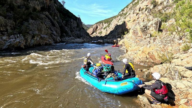 A small group of people wear helmets and board a raft at the edge of a river.