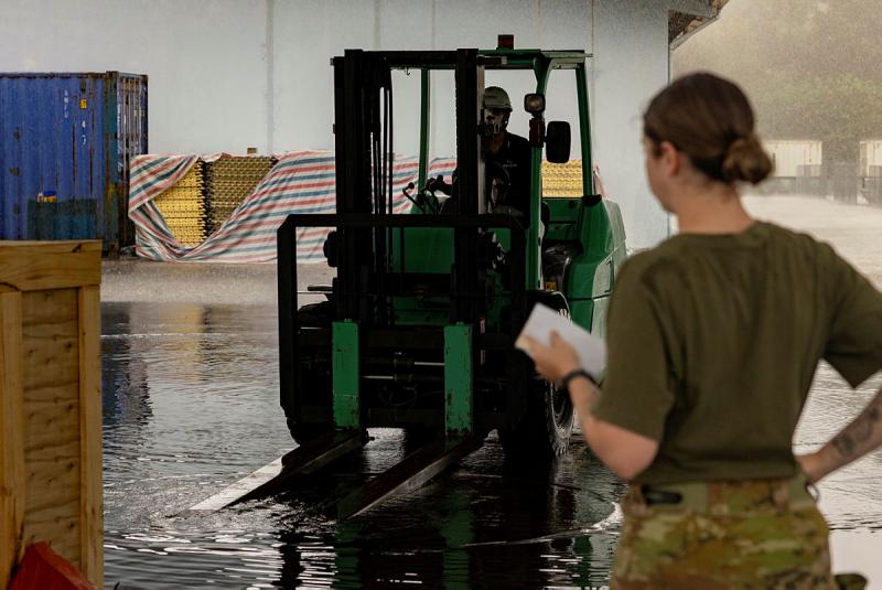A military woman oversees a man moving a small lifting vehicle.