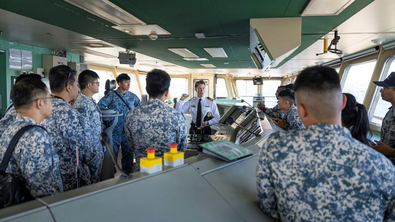 A person in a black and white Navy uniform speaks to a group of people in a different uniform inside a ship.