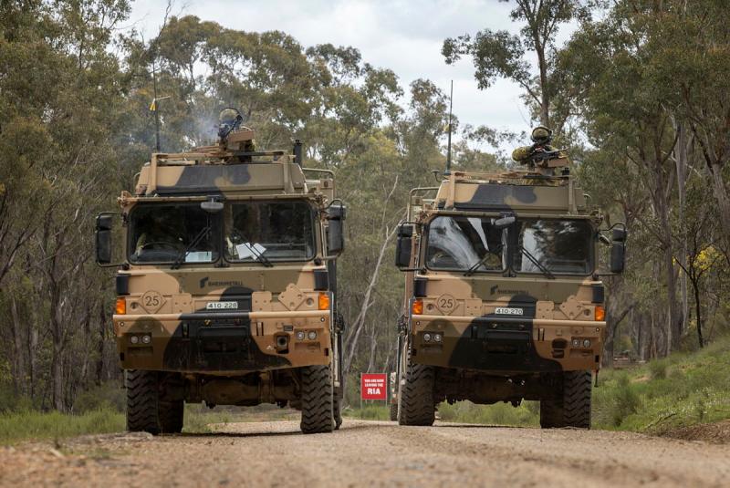 Two military vehicles drive down a bush road with people in uniform aiming weapons out of the vehicle roofs.