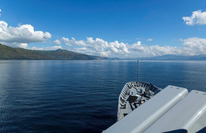 A ship’s bow pointed towards a green, hilly shoreline in the distance.