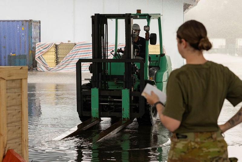 A military woman oversees a man drive a forklift through shallow water.