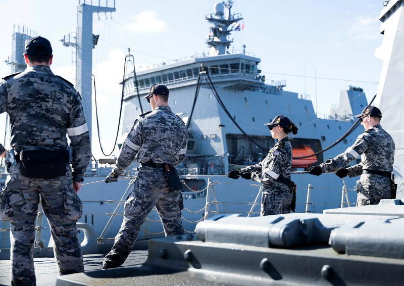 Four people stand near the rail of a ship, facing out towards another ship, three of them holding a line in both hands.