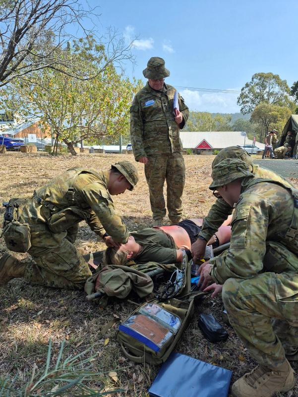 Army personnel standing and squatting around a person lying on a stretcher on the ground with burns makeup.