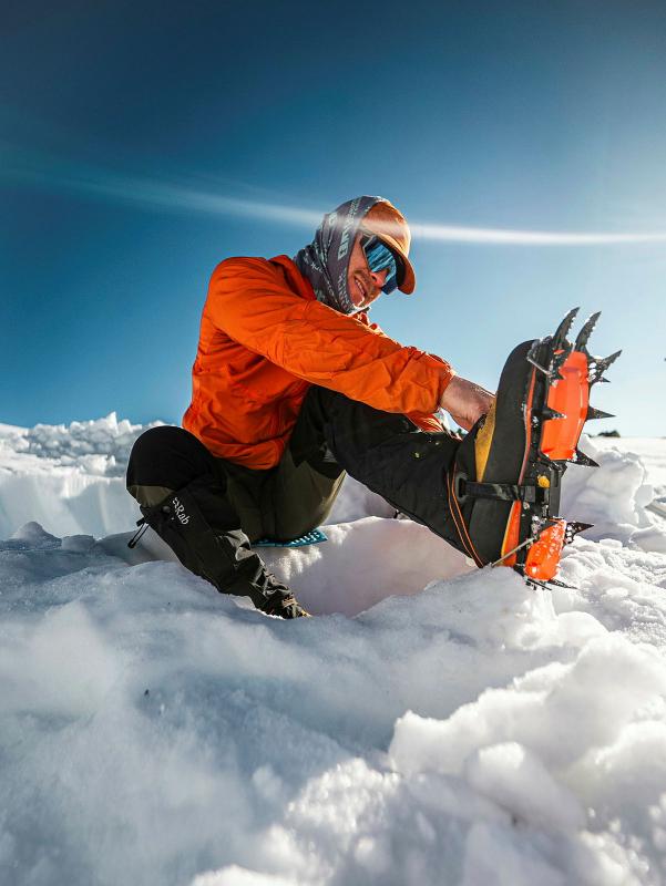 A hiker puts spikes on their boots while sitting in the snow.