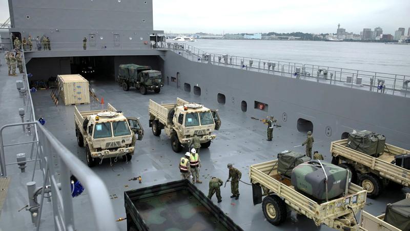 Military vehicles are parked on the deck of a ship, with some people in uniform working around them.