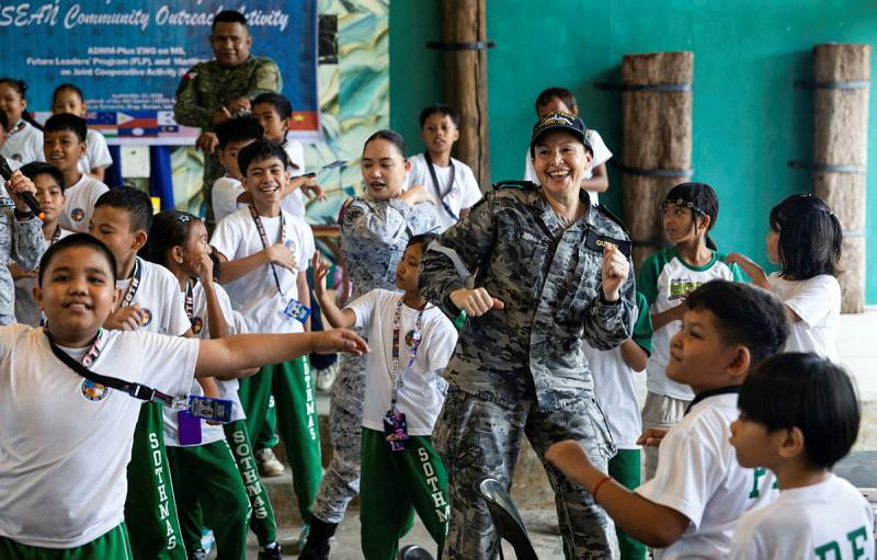 A military woman dances with a group of school children.