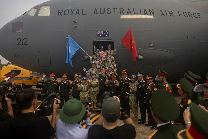 A group of military personnel stand in front of an aircraft with flags, while waving to a crowd.