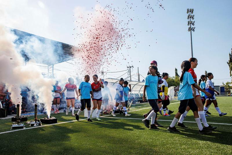 Soccer players and students walk hand in hand onto a field while confetti rains.