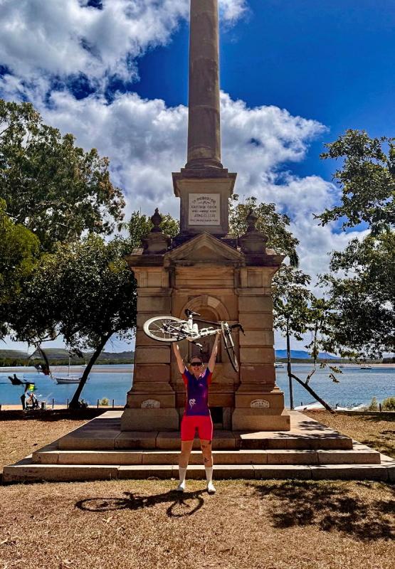 A woman holds a bicycle above her head in front of a memorial beside the waterfront.
