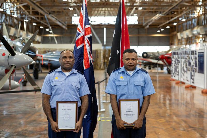 Two military men holding certificates stand side by side in front of two national flags and several stationary aircraft.