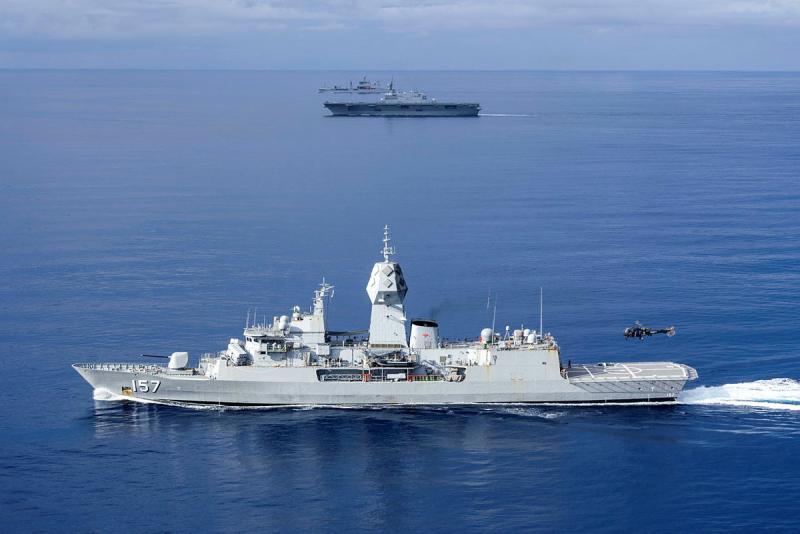 Three ships sail side by side while a helicopter hovers above the flight deck of one ship.