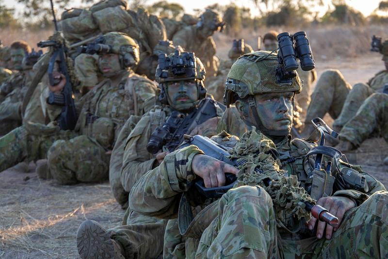 Army soldiers wearing full military kit sit together in a group on the dirt ground.