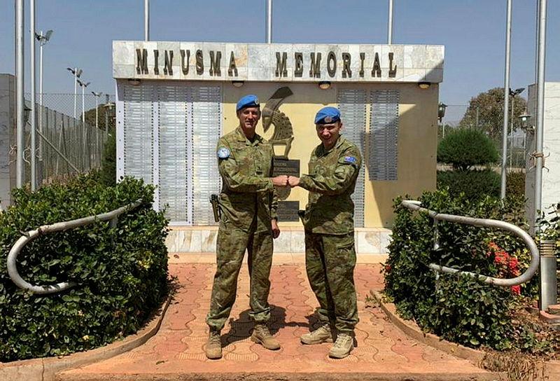 Two men in uniform and blue peacekeeping berets fist bump in front of a memorial.