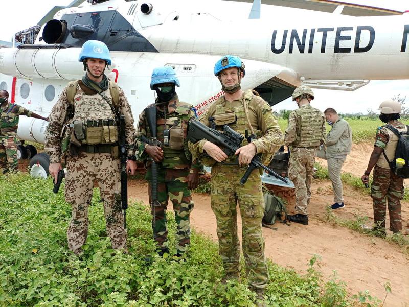 Three men in uniform and blue helmets hold weapons in front of a helicopter.