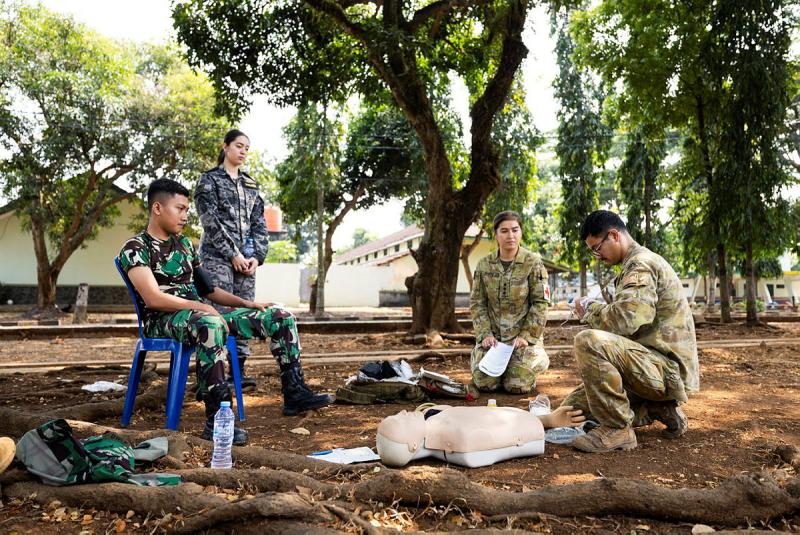 Military personnel in varied uniforms crowd around a 'casualty' mannequin in an outdoor setting.