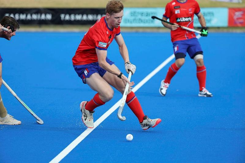 A man in a red and blue uniform plays field hockey on a blue pitch.