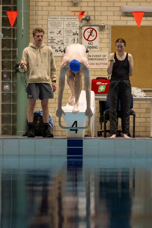 A man prepares to dive from a swim block while two people stand to either side of the block.