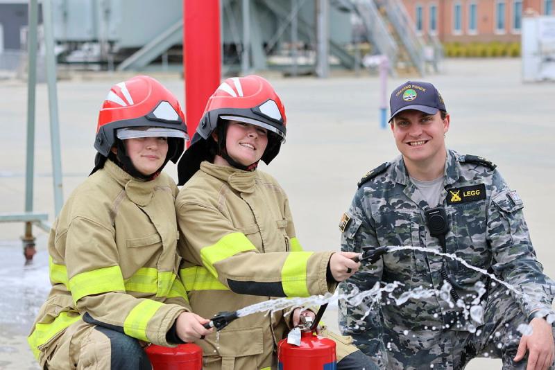 Two people in firefighting gear kneel while using fire extinguishers as a uniformed man kneels beside them.