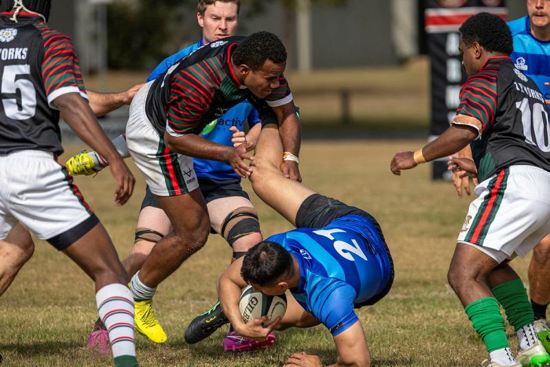 A rugby player carrying the ball falls to the ground as an opponent grabs his leg.