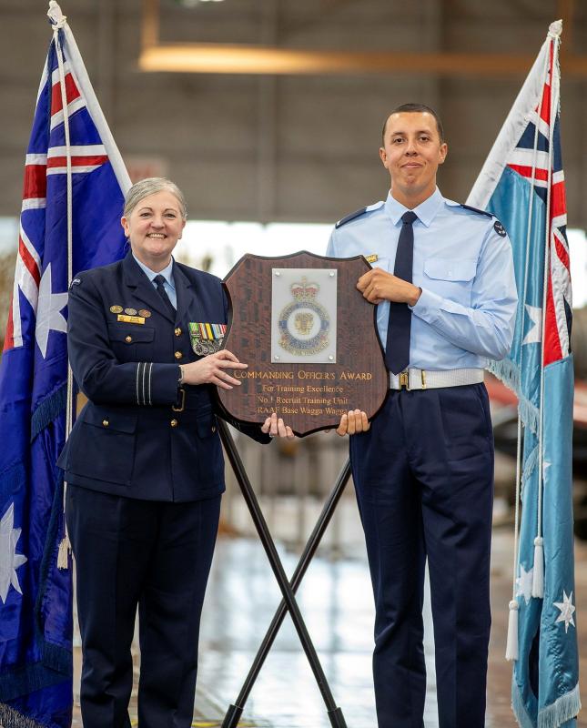 A military woman and man hold an award together, with national flags behind them.