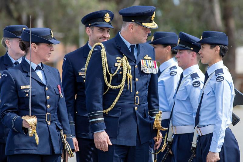 Men and women in dark blue military uniform walk past a row of young people in military uniform.