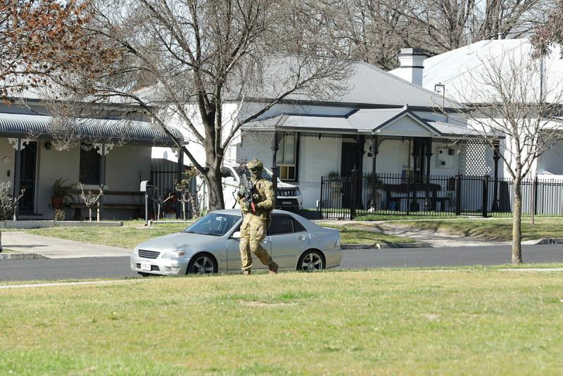 A soldier carrying a rifle walks along a suburban footpath with houses visible across the street.