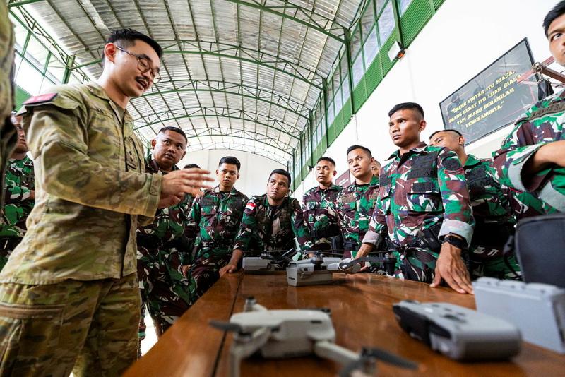 A group of military people in one type of uniform watch another military person in a different uniform speak with drones on a table nearby.