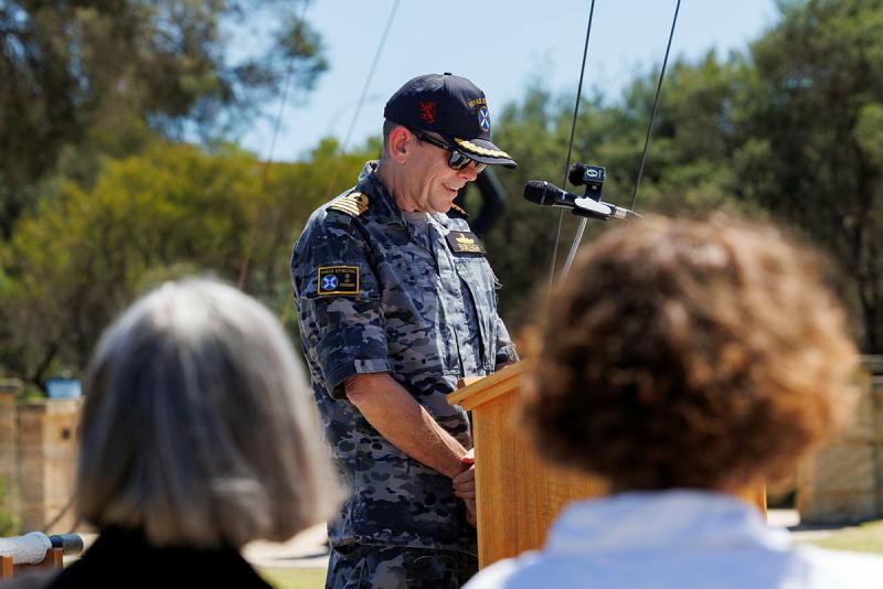 A man in a Navy uniform smiles and stands in front of a microphone outdoors.