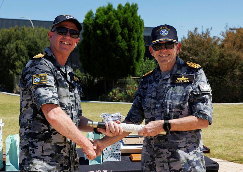 A man in a Navy uniform passes an object to another man in Navy uniform in an outdoor setting.