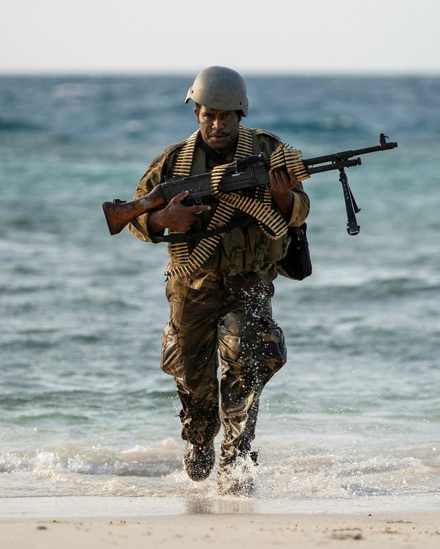 A man in uniform runs out of the water onto a beach holding a rifle, with belts of ammunition draped across his body.