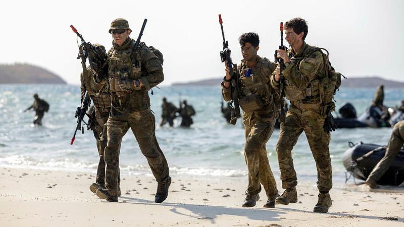 Four men in uniform run on a beach holding rifles, with others in the water and on boats behind them.