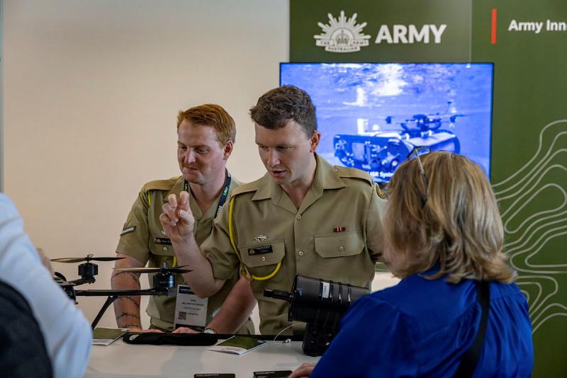 Two men in uniform stand behind a table with a drone on it, one talking to people standing on the other side of the table.