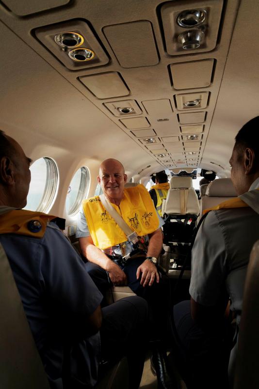 Man in a yellow life jacket seated in a narrow plane talks to two people sitting across from him