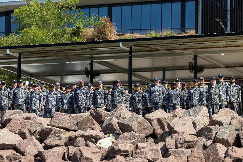 Rows of people in Navy uniform stand on the shore near a sea rock wall.