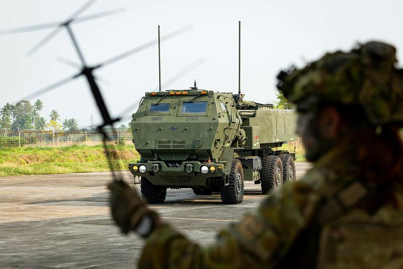 A large military vehicle sits on a concrete surface while a person in uniform is out of focus in the foreground.
