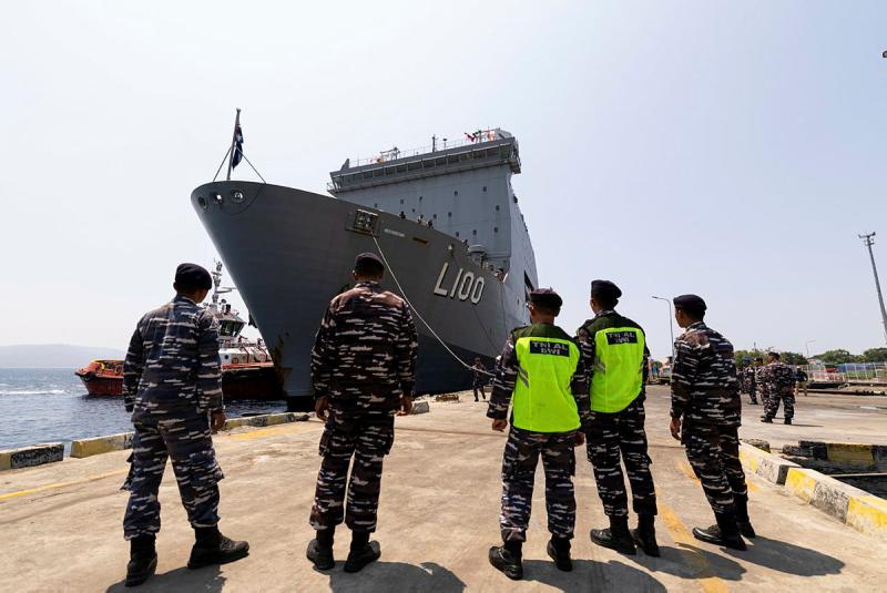 Military personnel stand on the shore as a ship comes into port.