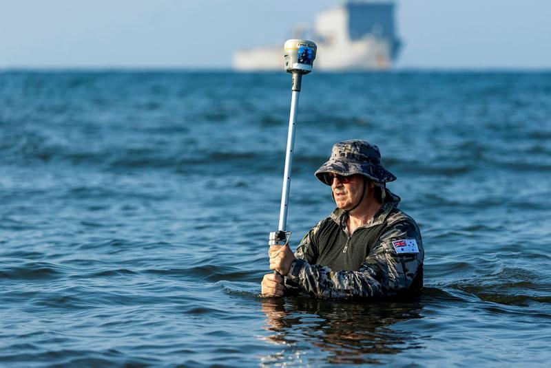A man in Navy uniform holding a metal rod while standing waist-deep in ocean water.