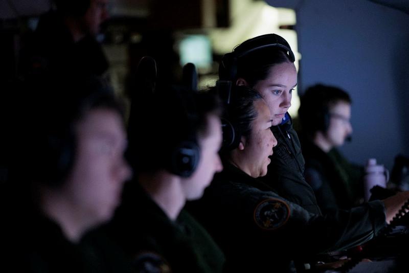 Five military personnel wear headphones and work in front of screens.
