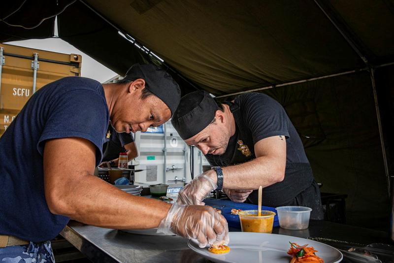 Two men lean over plates, placing food on them.
