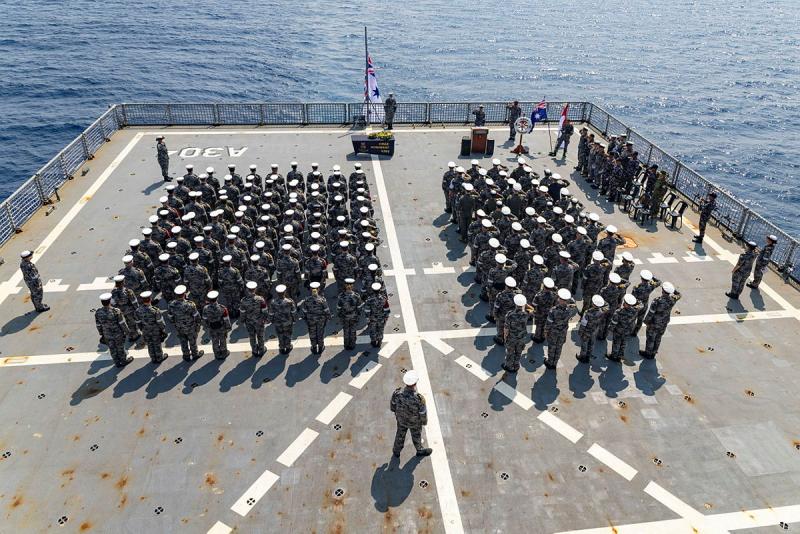 Numerous people in uniform stand in rows on the deck of a ship, facing out to sea, while a wreath lies on a table in front of them.