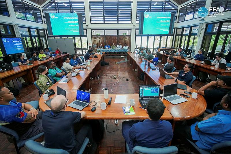 People sit at wooden desks arranged in a U formation with large screens in the background displaying text