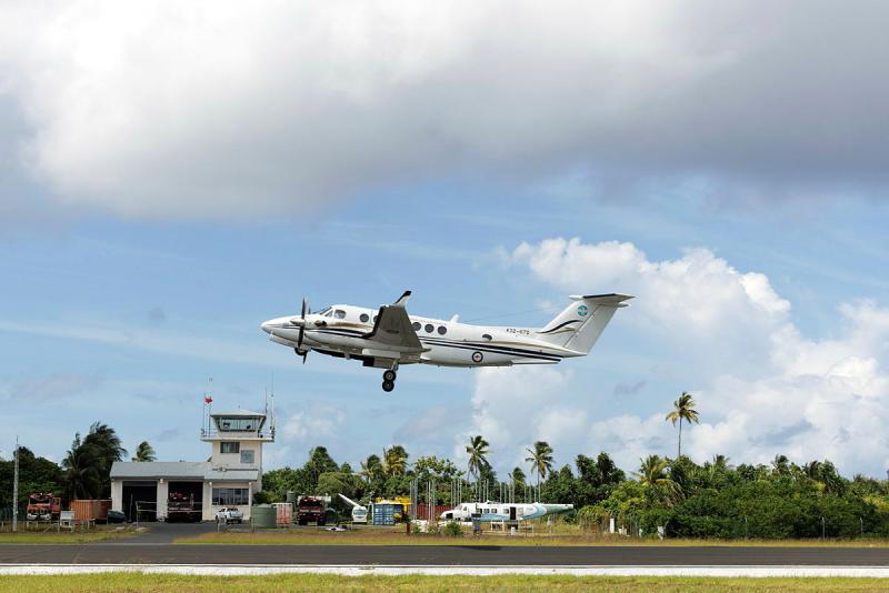 An aircraft takes off from a runway, palm trees and greenery on the ground below and behind it